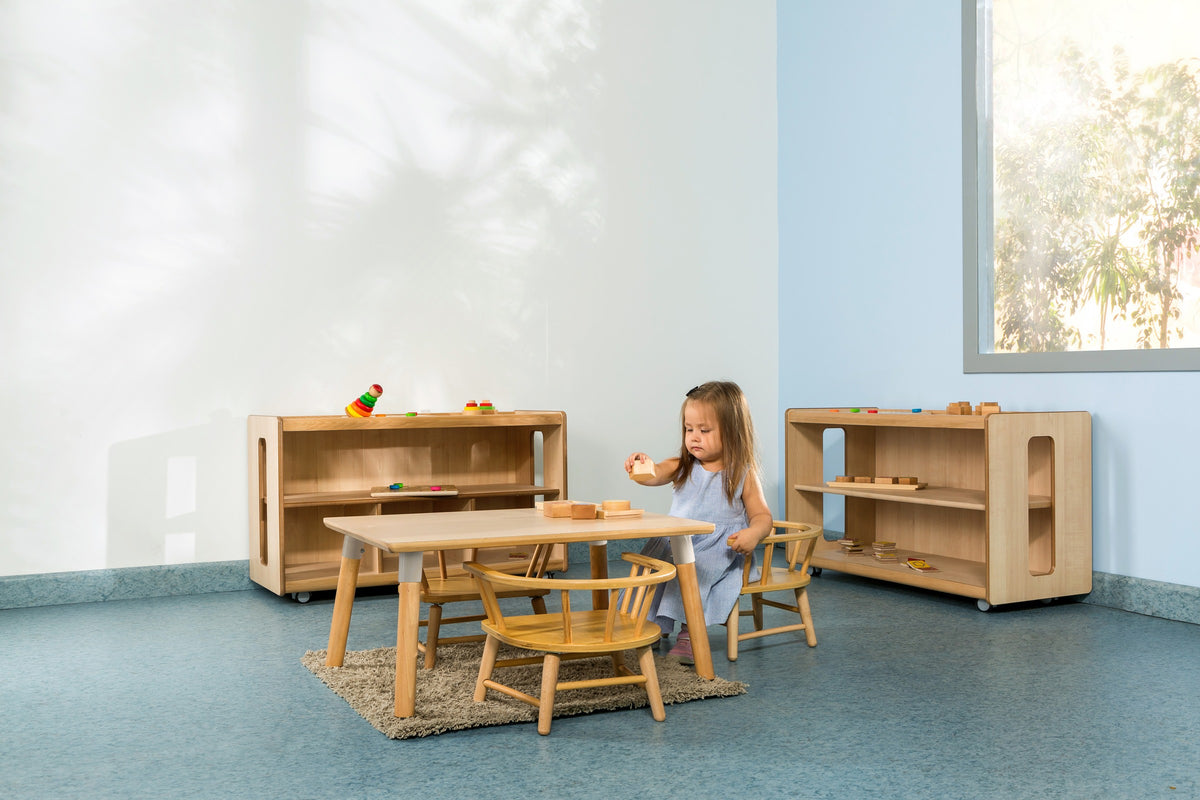 A child using a low timber table and curved armchair beside open shelving.