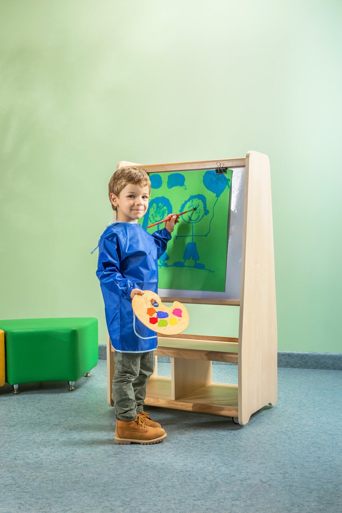 A child painting at a wooden easel with paint pots on its lower shelf.